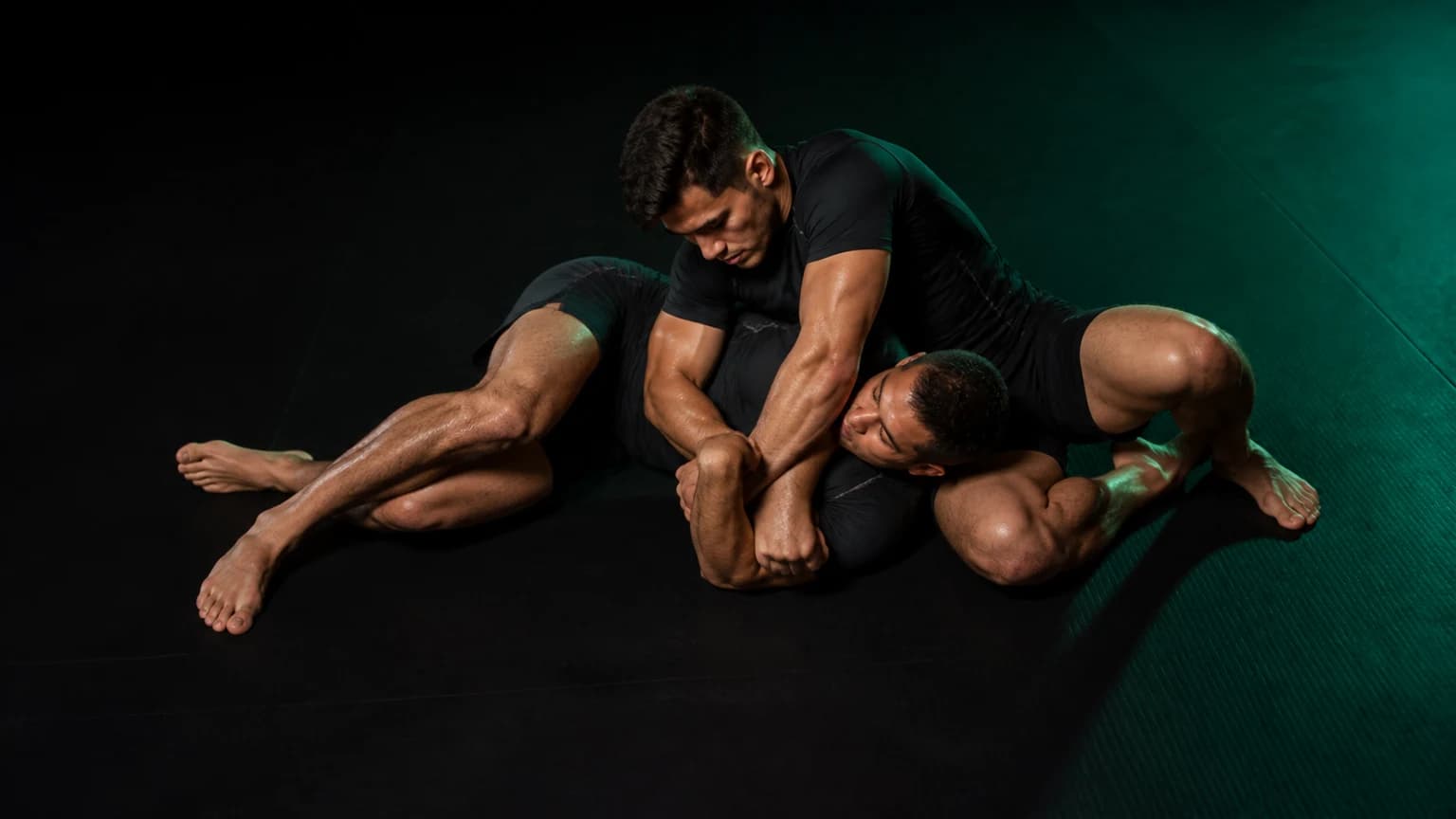 Two grapplers demonstrate the arm triangle finishing position slowly on an emerald and charcoal mat